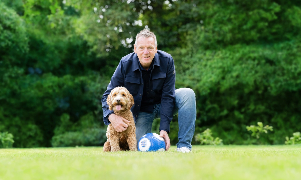 A man kneeling on grass beside a brown curly dog. He is holding a football on the ground.