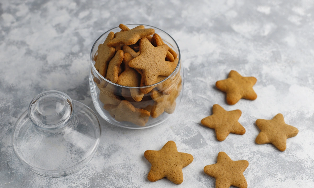 A glass jar filled with star-shaped gingerbread dog biscuits is open on a textured grey surface, with several biscuits scattered around it.