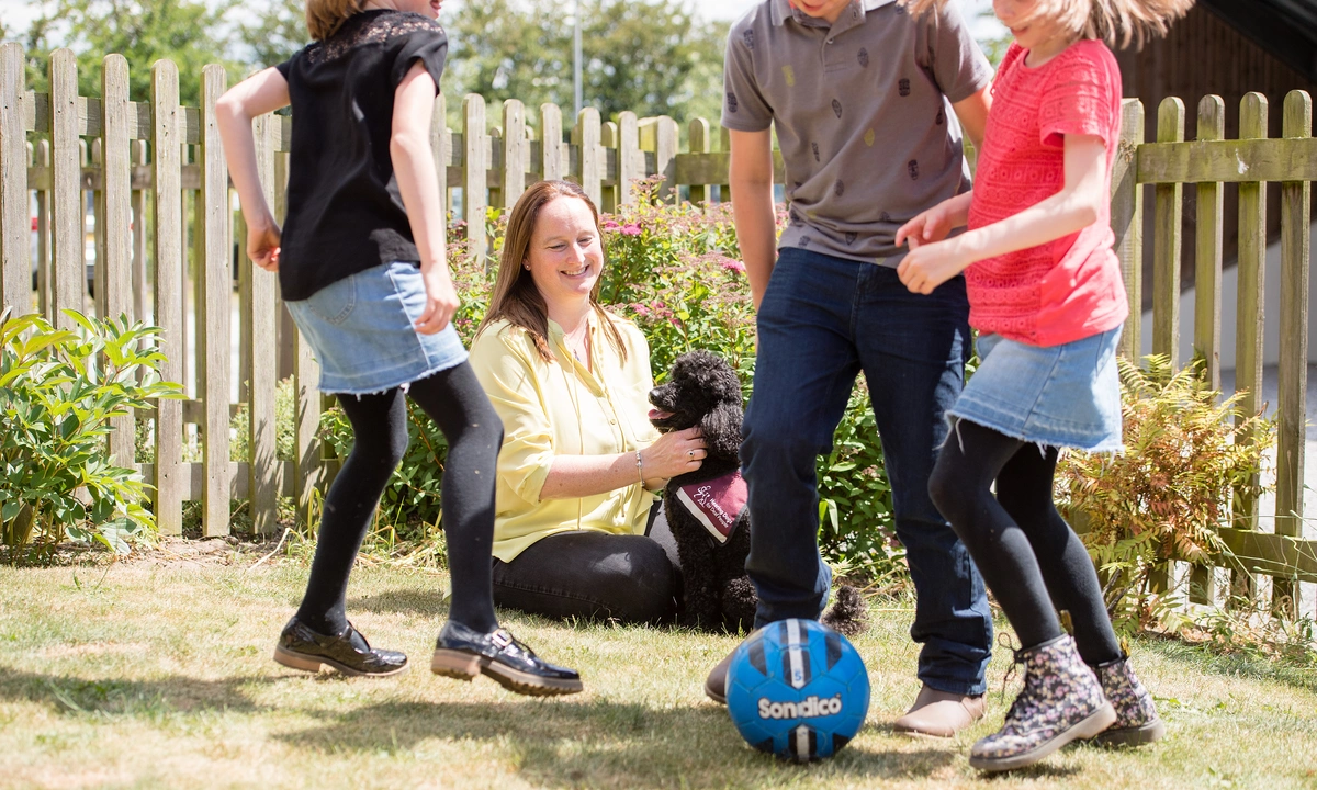 A woman is sat on grass in sunshine with her hearing dog poodle watching her three children playing football nearby