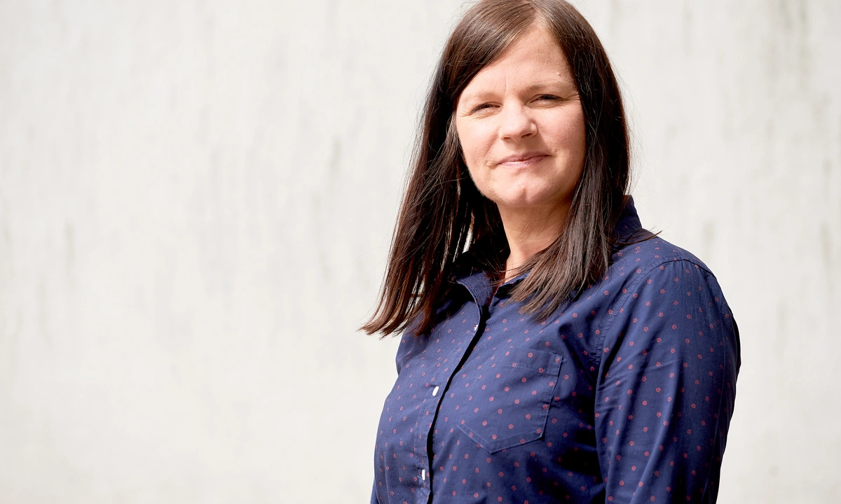 Mid shot of a woman with long brown hair and a dark blue shirt with a light grey textured background