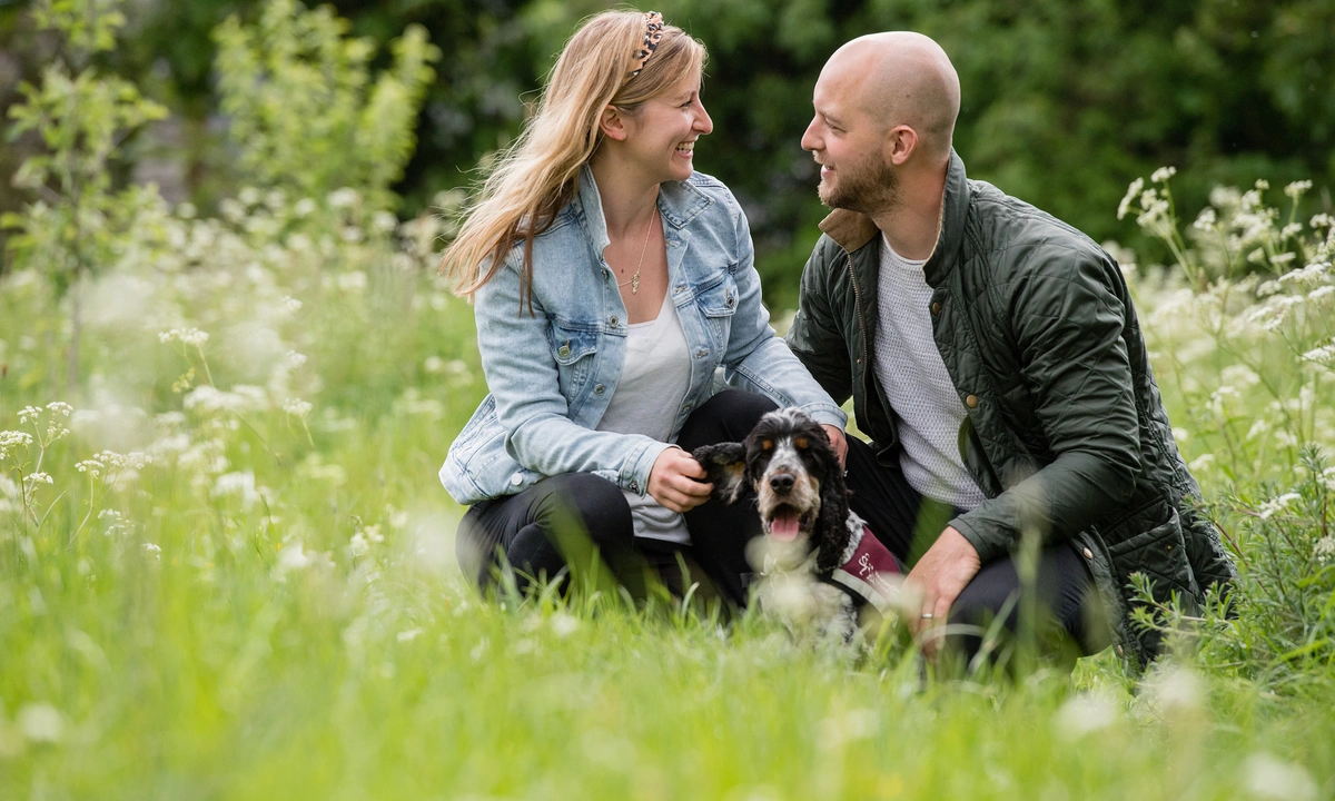A man and a woman looking at each other in a field of flowers with a hearing dog Spaniel sat in front of them