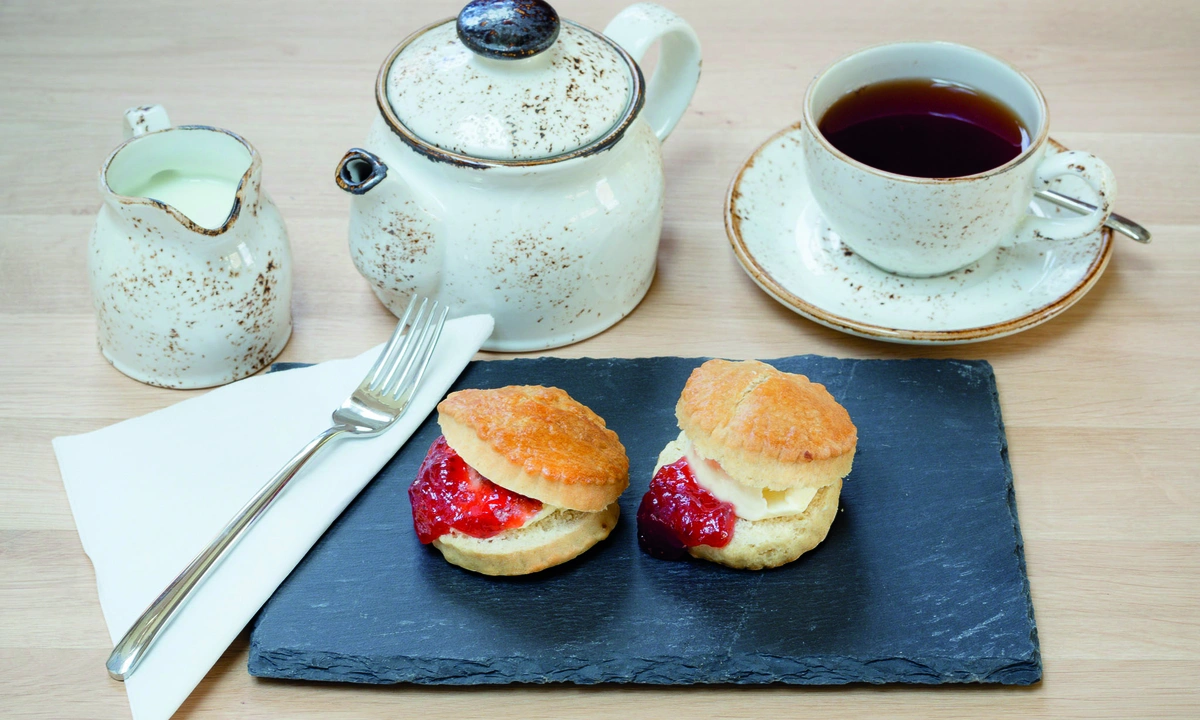 Teapot with cup of tea and milk, and two scones infront next to a fork and napkin