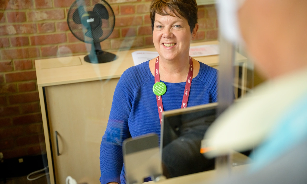 Smiling woman sat at a reception desk wearing a staff lanyard and a lipreading badge greeting a person