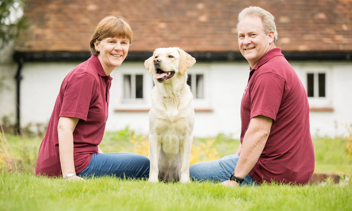 A happy white labrador sitting on grass between a man and a woman in hearing dogs volunteer uniform.