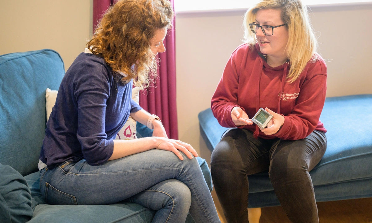 Two women sat in a lounge, the one is hearing dogs uniform is showing the other how to use a training timer