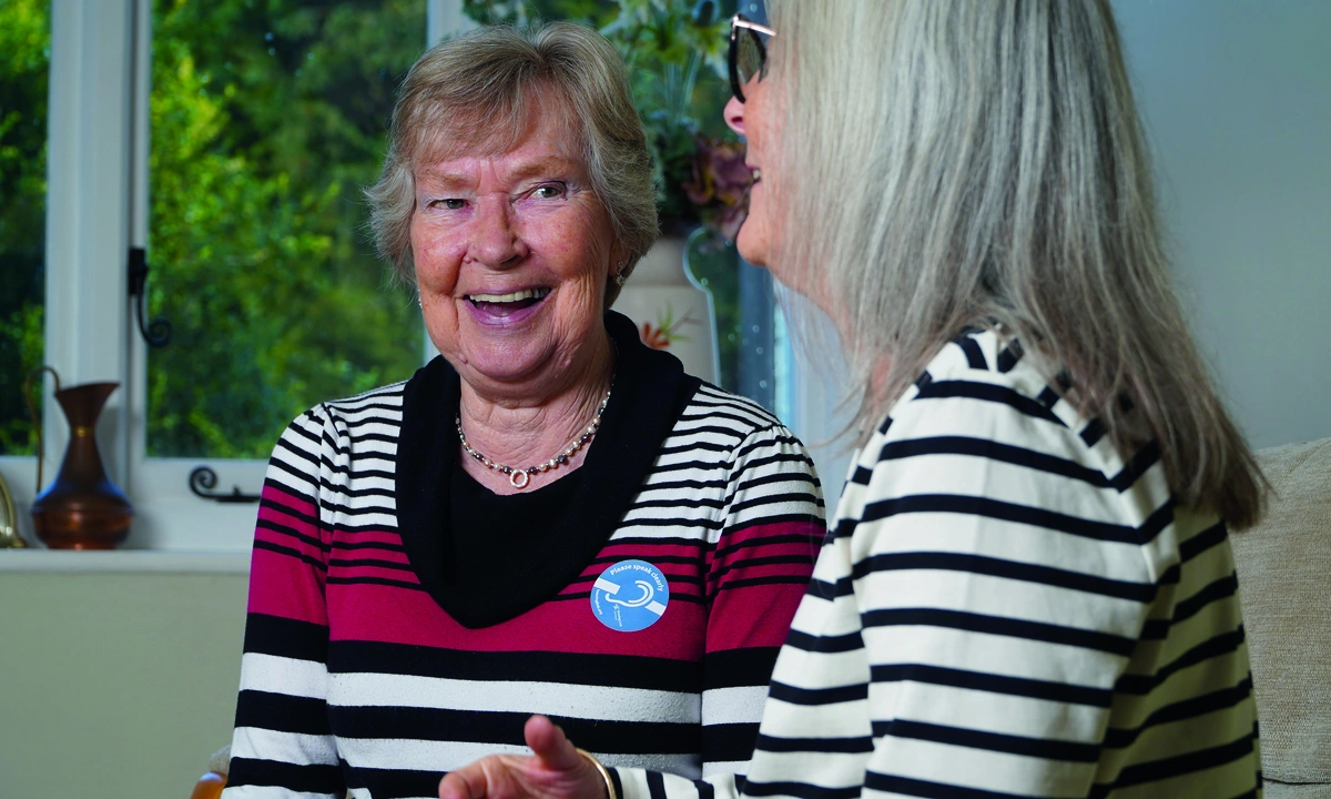 Two older women talking and smiling, one if wearing a badge with a deaf symbol that says ‘Please speak clearly’