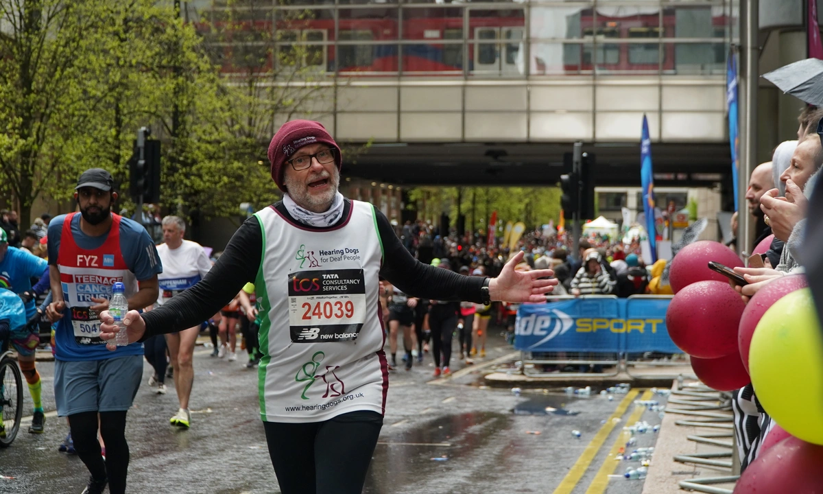 Runner called Martin in Hearing Dogs for Deaf People top and burgundy beanie hat running with is arms wide looking at the camera with a bridge and lots of other people at Great North Run