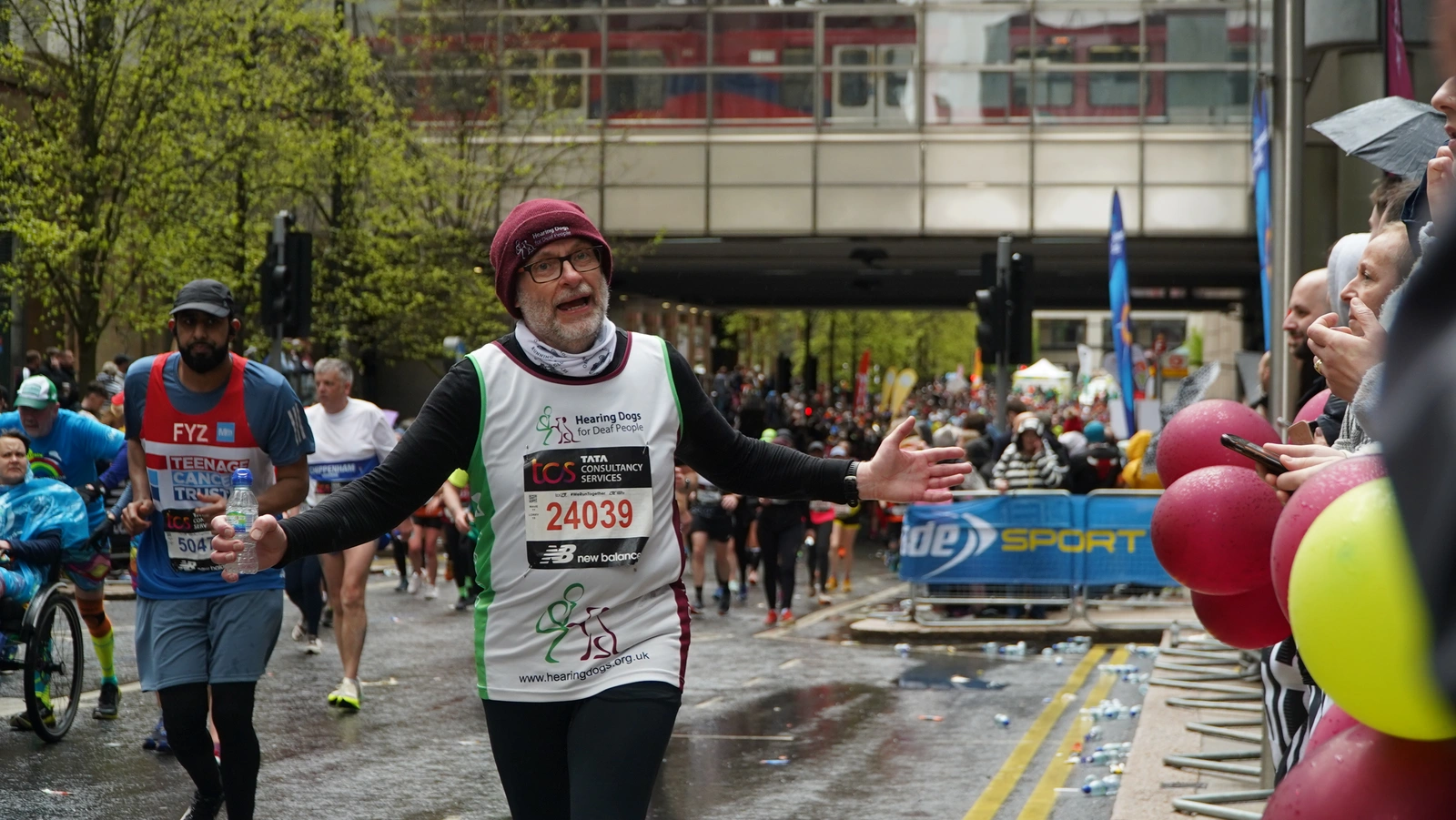 Runner called Martin in Hearing Dogs for Deaf People top and burgundy beanie hat running with is arms wide looking at the camera with a bridge and lots of other people at Great North Run