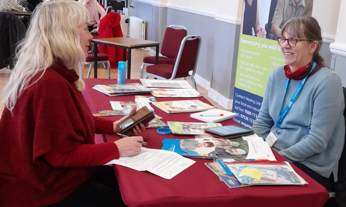 One woman talking to another one while sat at a table with literature on it