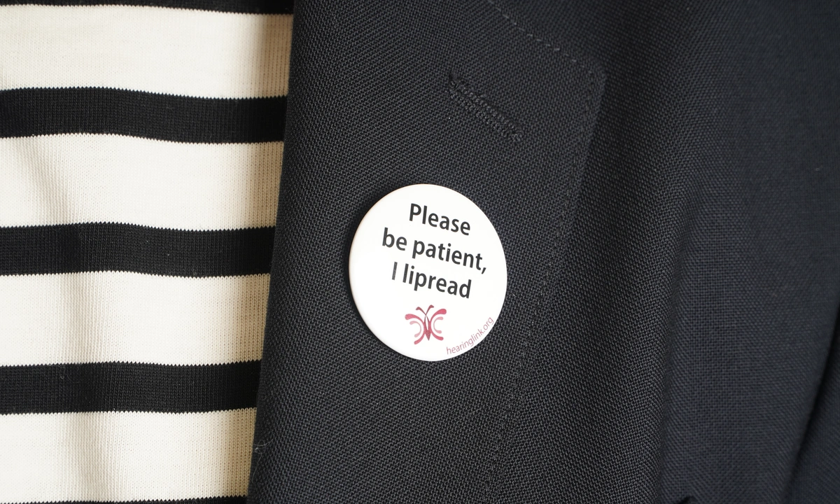 Close up of a badge on a jacket lapel that says ‘Please be patient, I lipread’
