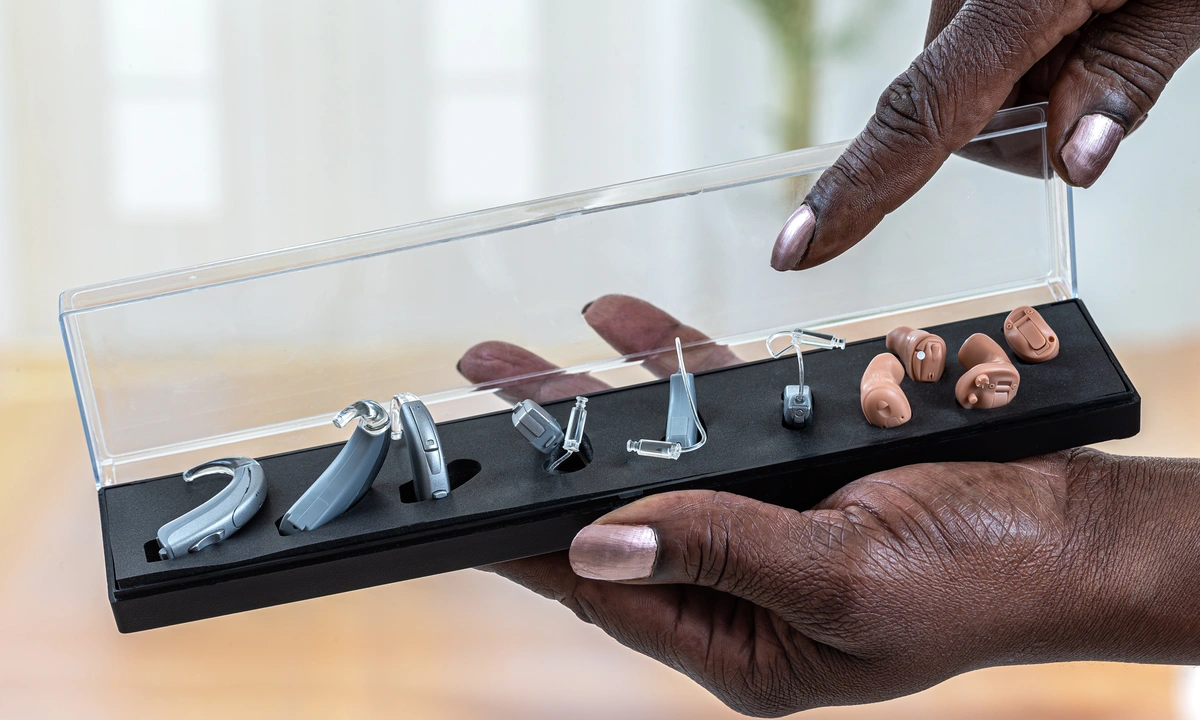 Close up of one hand holding a display case of four different types of hearing aid while the other hand points at them