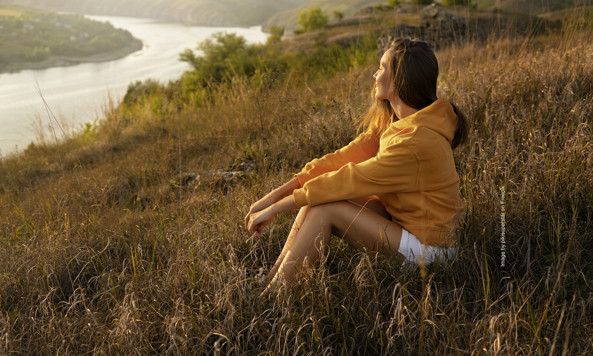 A woman sat on a hill as the sunsets looking out at some countryside