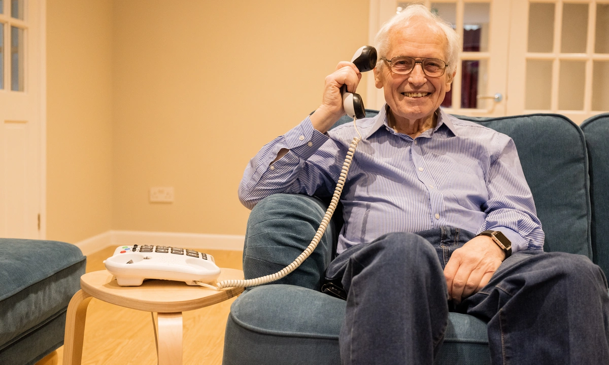 A man smiling sat on a sofa holding up a landline phone receiver to his ear