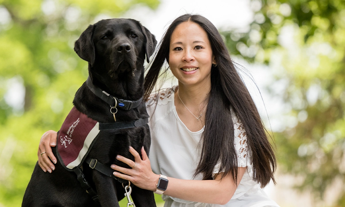 Mid shot of a smiling woman sat next to a black labrador with her arm around it, the dog is wearing a hearing dogs jacket