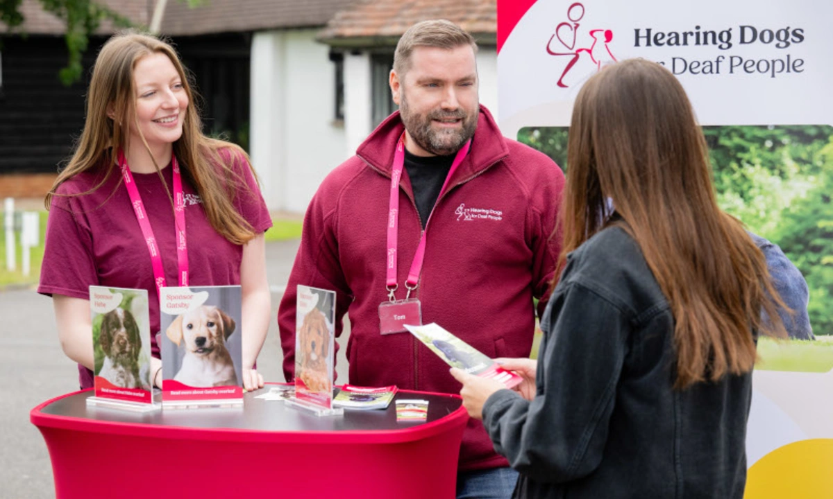 A man and 2 women are standing around a red table with brochures and pamphlets on it. Behind them is a banner that reads "Hearing Dogs for Deaf People"