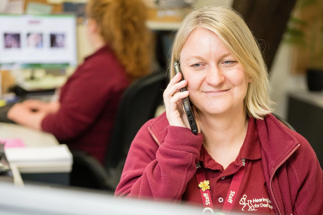 Woman, smiling, in hearing dogs uniform sat in an office in front of a laptop on the telephone 