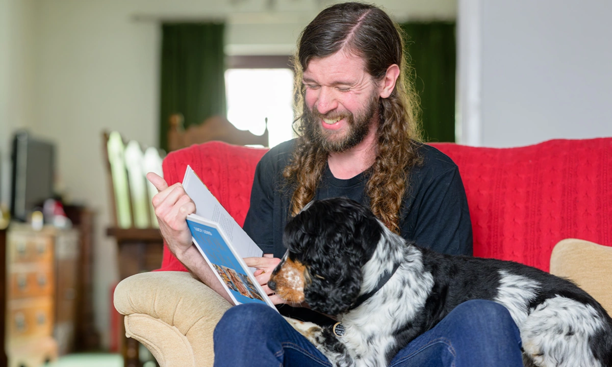 A man sat on a sofa reading a book and looking happy while his spaniel sits across his lap