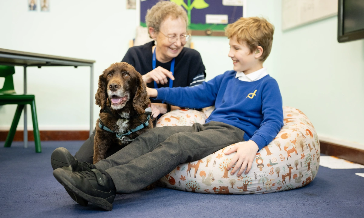 A young boy in blue uniform jumper is seated on a bean bag chair. He is stroking a brown down as a woman kneels smiling beside them