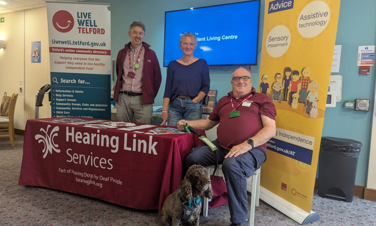 Three people and a dog assembled around a display table. A man and woman standing behind the table and a man sitting at the end holding the dog's lead