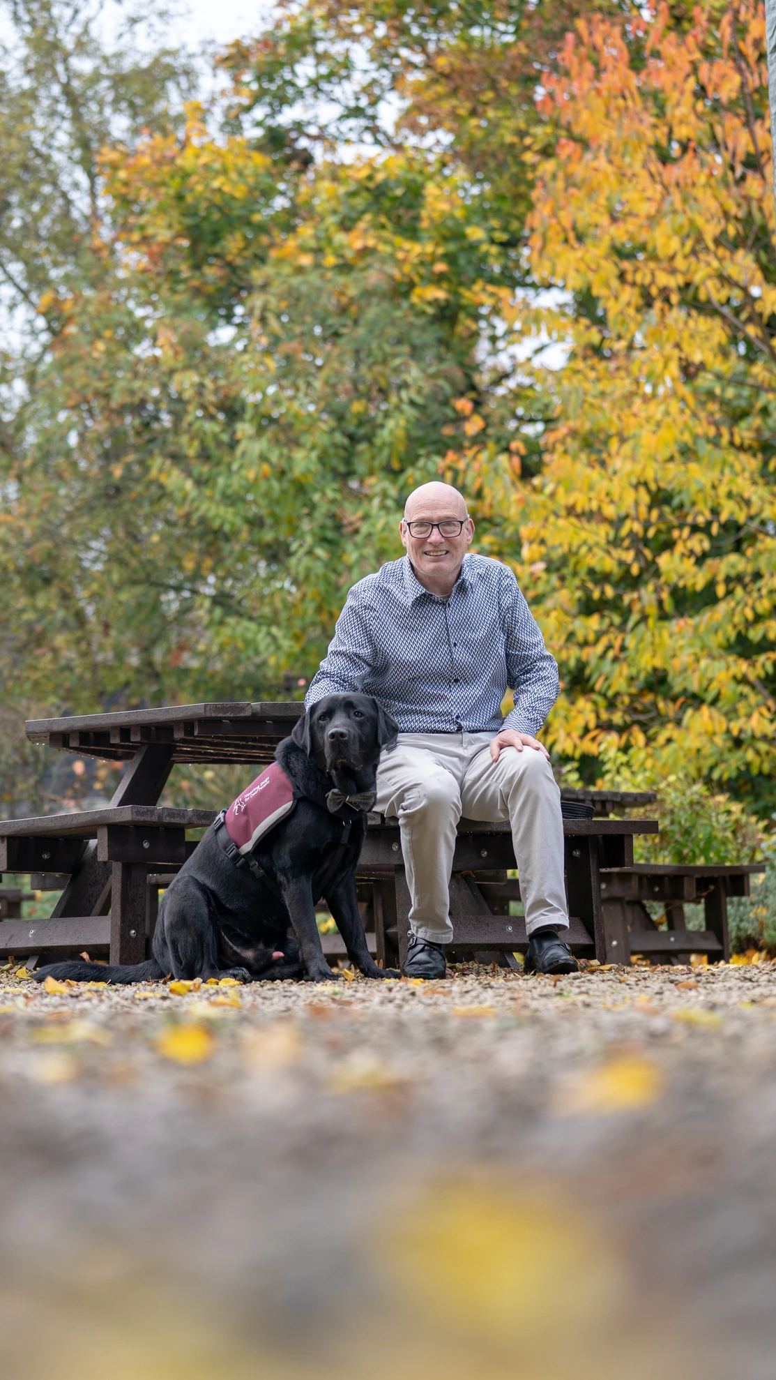 A man is sitting at a picnic table with a black labrador which is wearing a hearing dogs burgundy jacket. Trees with autumn coloured leaves are in the background.