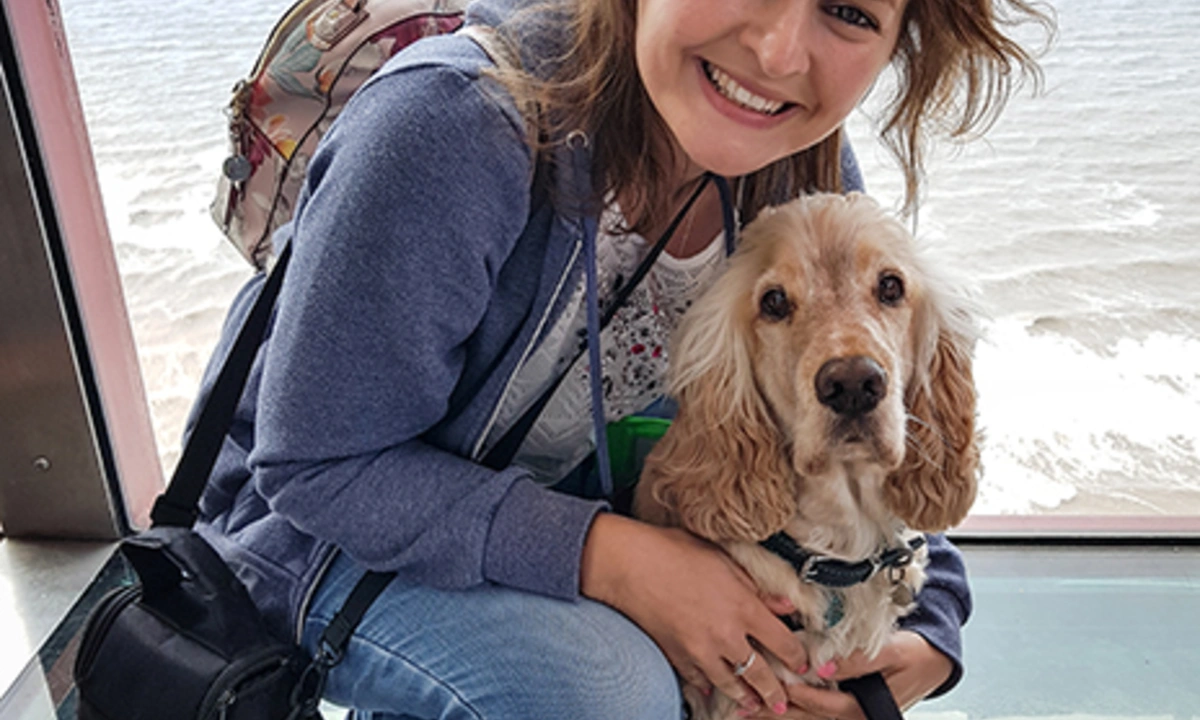 A smiling woman kneeling and hugging a light haired dog. They appear to be high above ground inside a structure with windowed walls and floor. A body of water can be seen behind them.