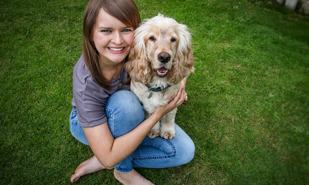 A young woman and light coloured dog are sitting on grass. The woman is smiling and hugging the dog.