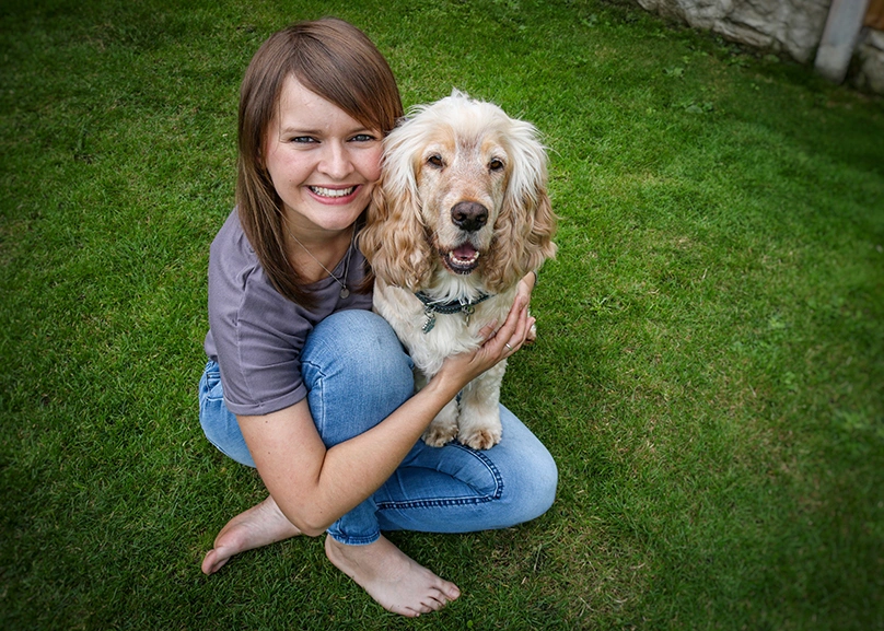 A young woman and light coloured dog are sitting on grass. The woman is smiling and hugging the dog.