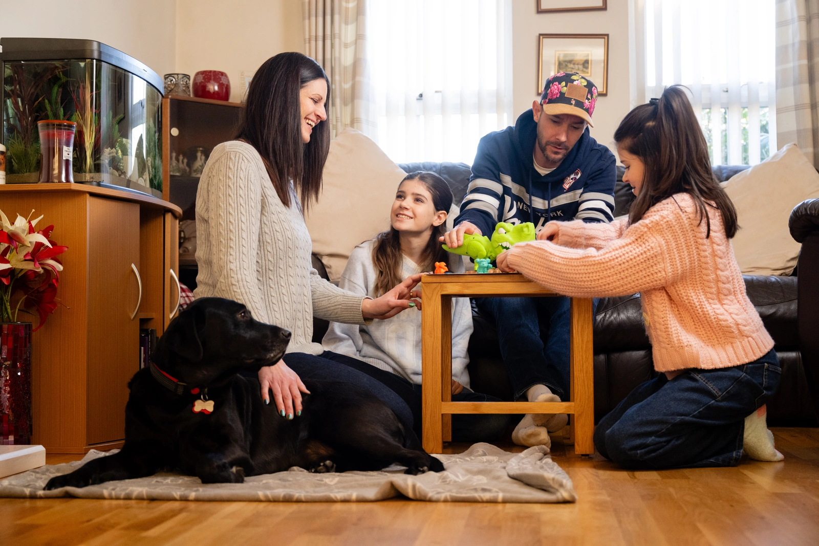 A family of four and a black labrador are sitting around a small table in a living room. The mood is happy and playful.