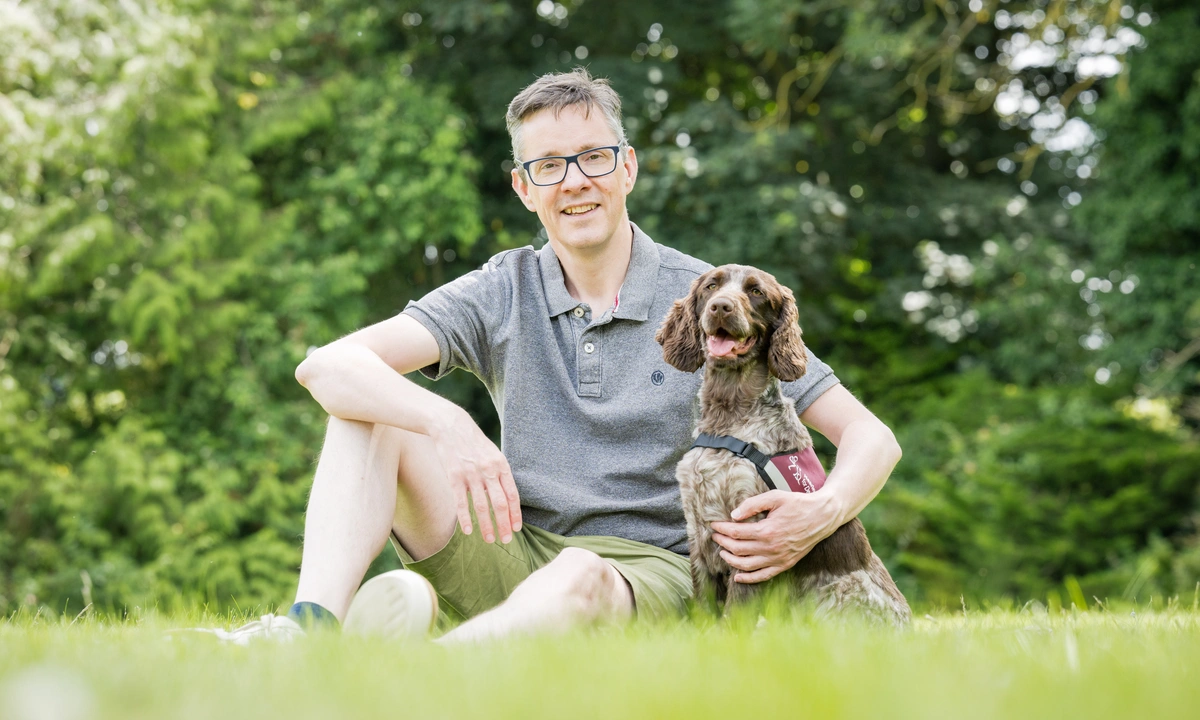 A smiling man, wearing spectacles, sitting on grass, with trees visible behind him. His left arm is around a brown and white cocker spaniel, which is wearing a hearing dogs jacket.