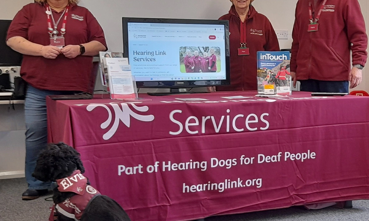 2 women and a man standing behind a table draped in a burgundy cloth. A black hearing dog sits in front.