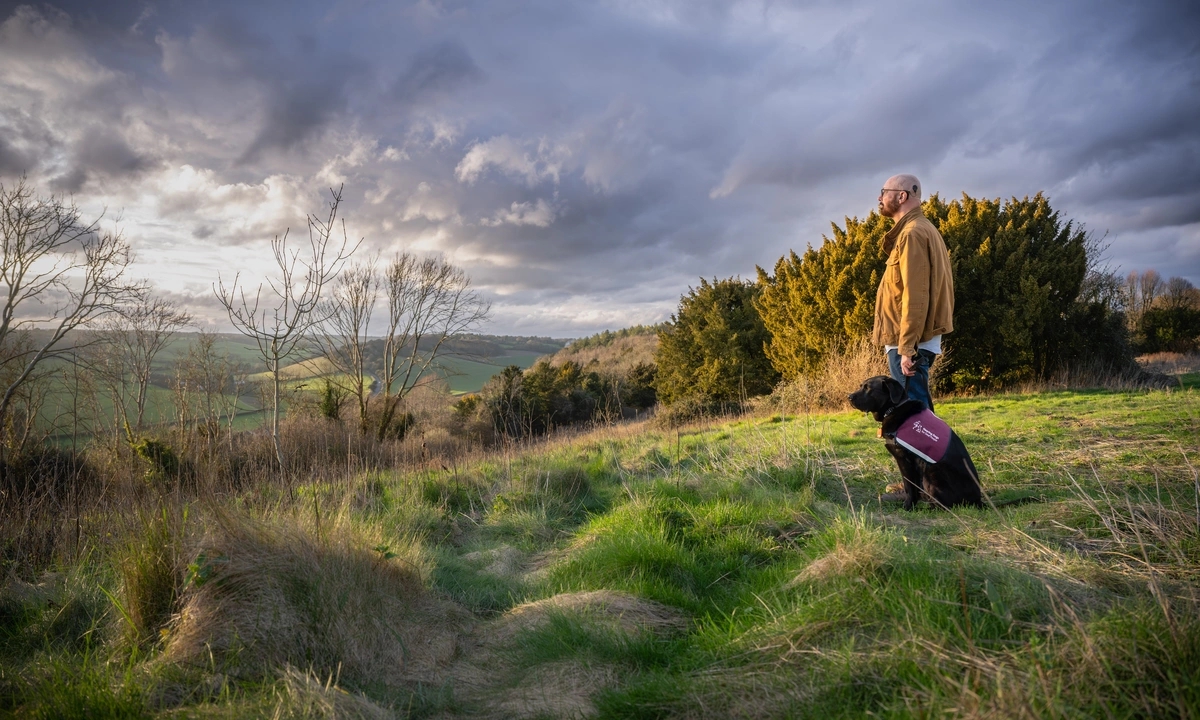 A photograph of Trevor and a black hearing dog gazing out across a dramatic outdoor scene of wild grass, green hills and clouds.