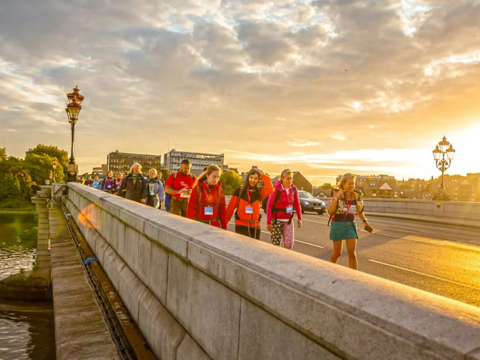 A photo showing a group of people walking across a bridge, the sky has dramatic clouds and the sun lights the right side of the image in a warm glow.