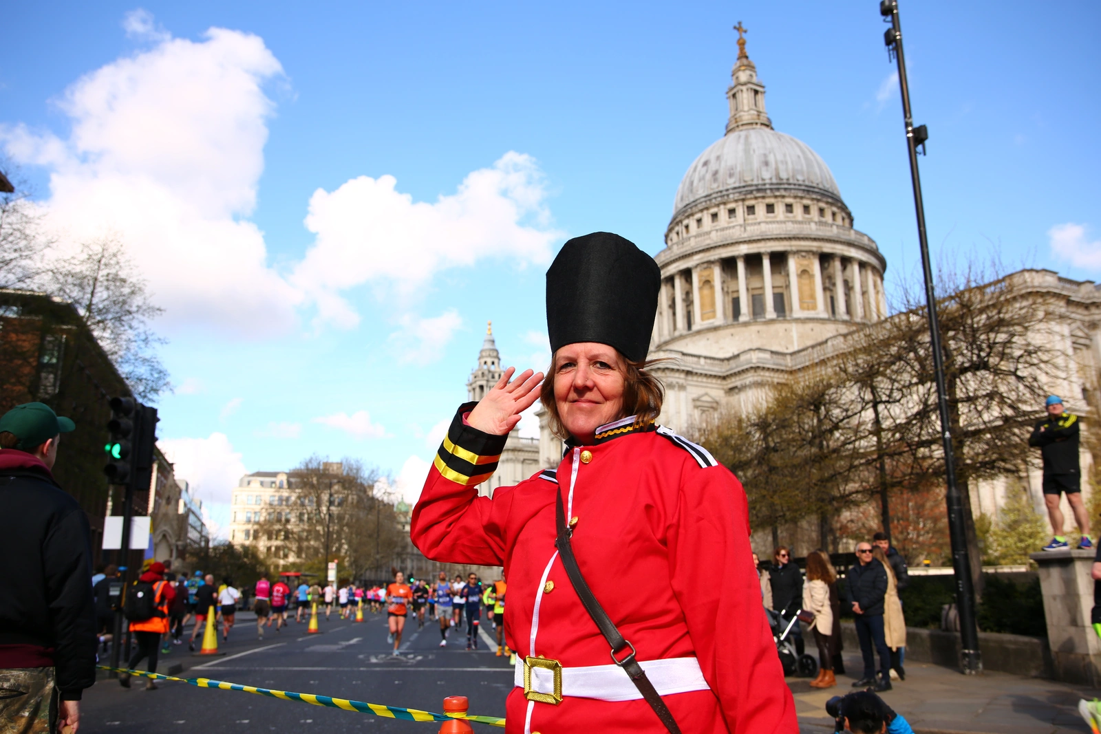 A woman, dressed in a mock Palace Guard costume, saluting. Behind her, marathon runners can be seen running past St. Paul's Cathedral.