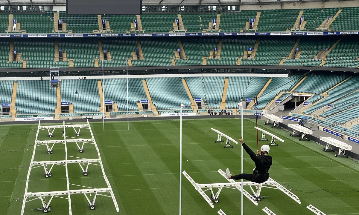 A view from high up in a large sports stadium, of a woman in black, ziplining across the pitch.