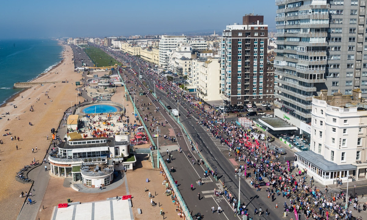 A birds eye view of hundreds of runners on the road alongside Brighton seafront. The beach and sea can be seen on the left, hotels on the right.