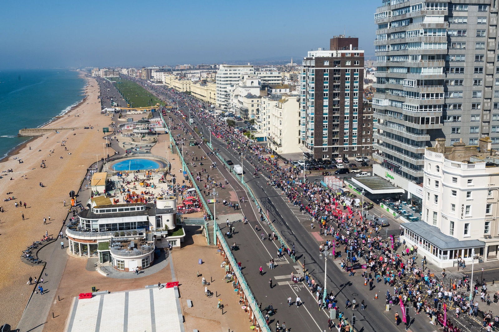 A birds eye view of hundreds of runners on the road alongside Brighton seafront. The beach and sea can be seen on the left, hotels on the right.