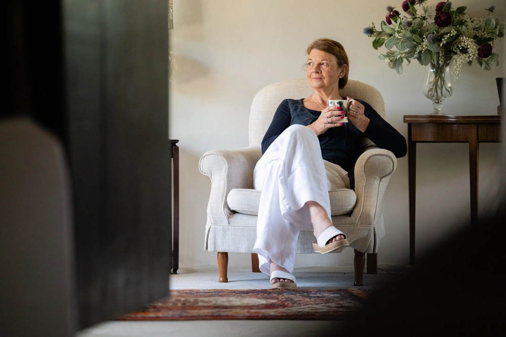 A woman sitting on a chair, holding a mug. She is looking off to the side contemplatively. 