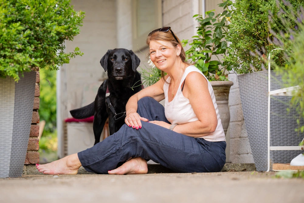A woman, Karen, is sitting on the ground in her garden. Behind her is a black labrador looking intently at camera.