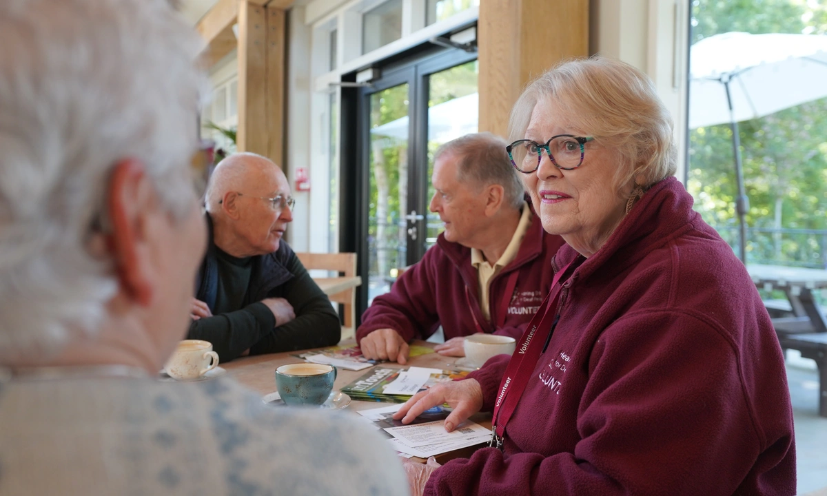 Seen over a woman's shoulder, a man and woman wearing hearing dogs volunteer uniforms are chatting to the other people at a table.