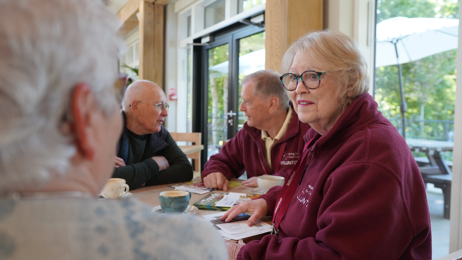 Seen over a woman's shoulder, a man and woman wearing hearing dogs volunteer uniforms are chatting to the other people at a table.