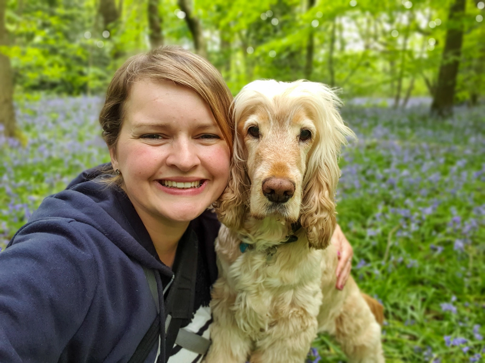 A smiling young woman and a light coloured dog in a in a woodland with blue flowers on the ground.