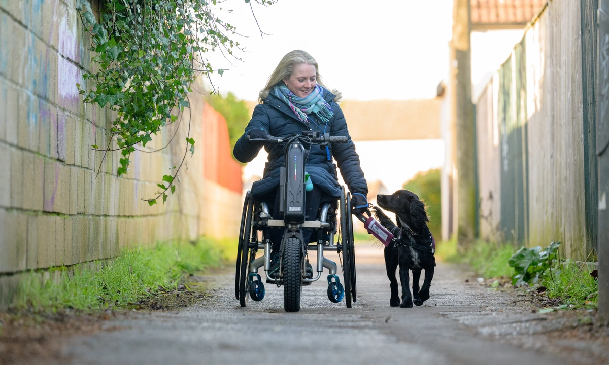 A smiling woman in a wheelchair, with a black dog walking alongside. They are looking at each other