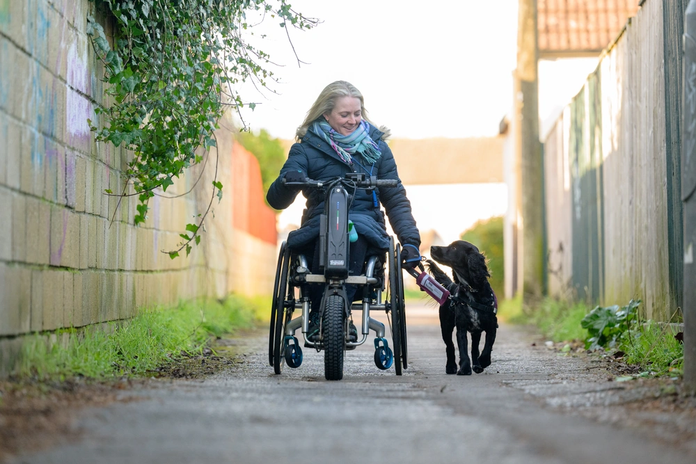 A smiling woman in a wheelchair, with a black dog walking alongside. They are looking at each other
