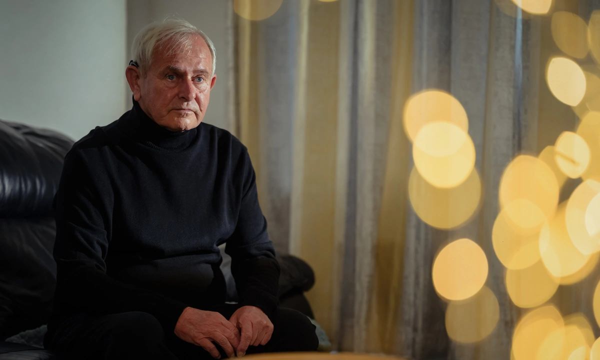 A saddened older gentleman, dressed in dark clothing sitting on a sofa. Blurry, out of focus fairy lights are in the foreground