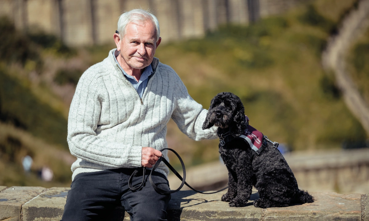 David sitting on a wall, with black cockapoo Asti sitting on top of the wall beside. Asti is wearing his hearing dogs jacket.