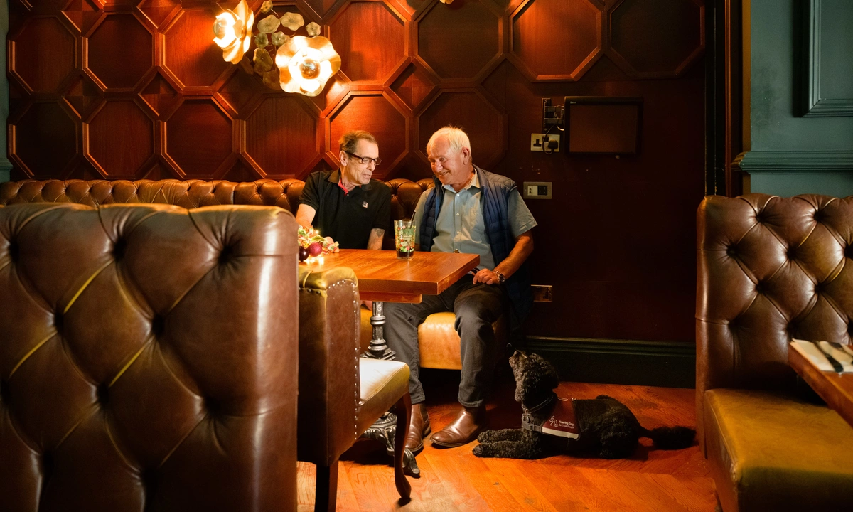David and a friend sitting at a table in a cosy restaurant with plush leather seats. Asti is settled on the floor beside David.