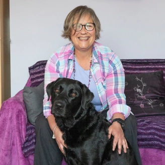 A woman sitting on a sofa. A black labrador dog is in front of her