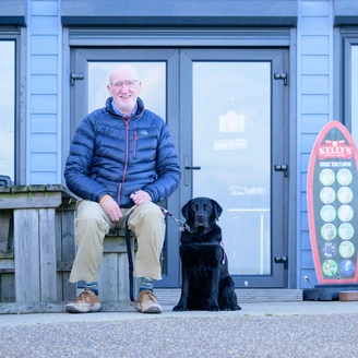A man sat outside a blue painted cafe smiling with a labrador sat next to hi