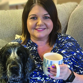 A smiling woman with brown, shoulder length hair is sitting on a sofa with her right arm is around a cocker spaniel dog