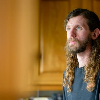Head and shoulders image of a young man with long brown hair looking into the distance with a sad expression